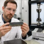 A materials scientist in a lab examines a carbon fiber reinforced composite sample near a tensile testing machine.